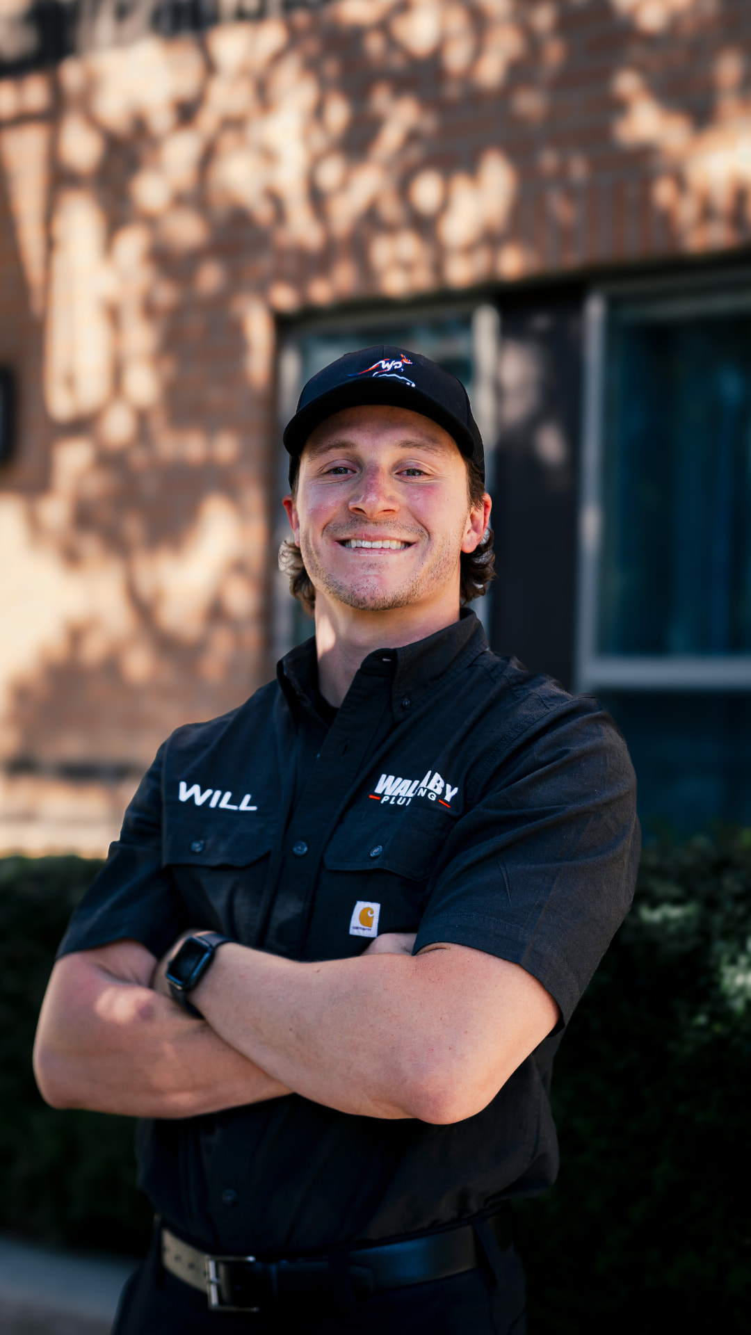 Will Pollet, owner of Wallaby Plumbing, smiling with arms crossed in a branded shirt and cap